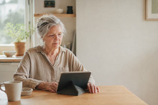 Image d’illustration : une femme âgée, assise à la table de sa cuisine, travaille sur une tablette posée sur un support.